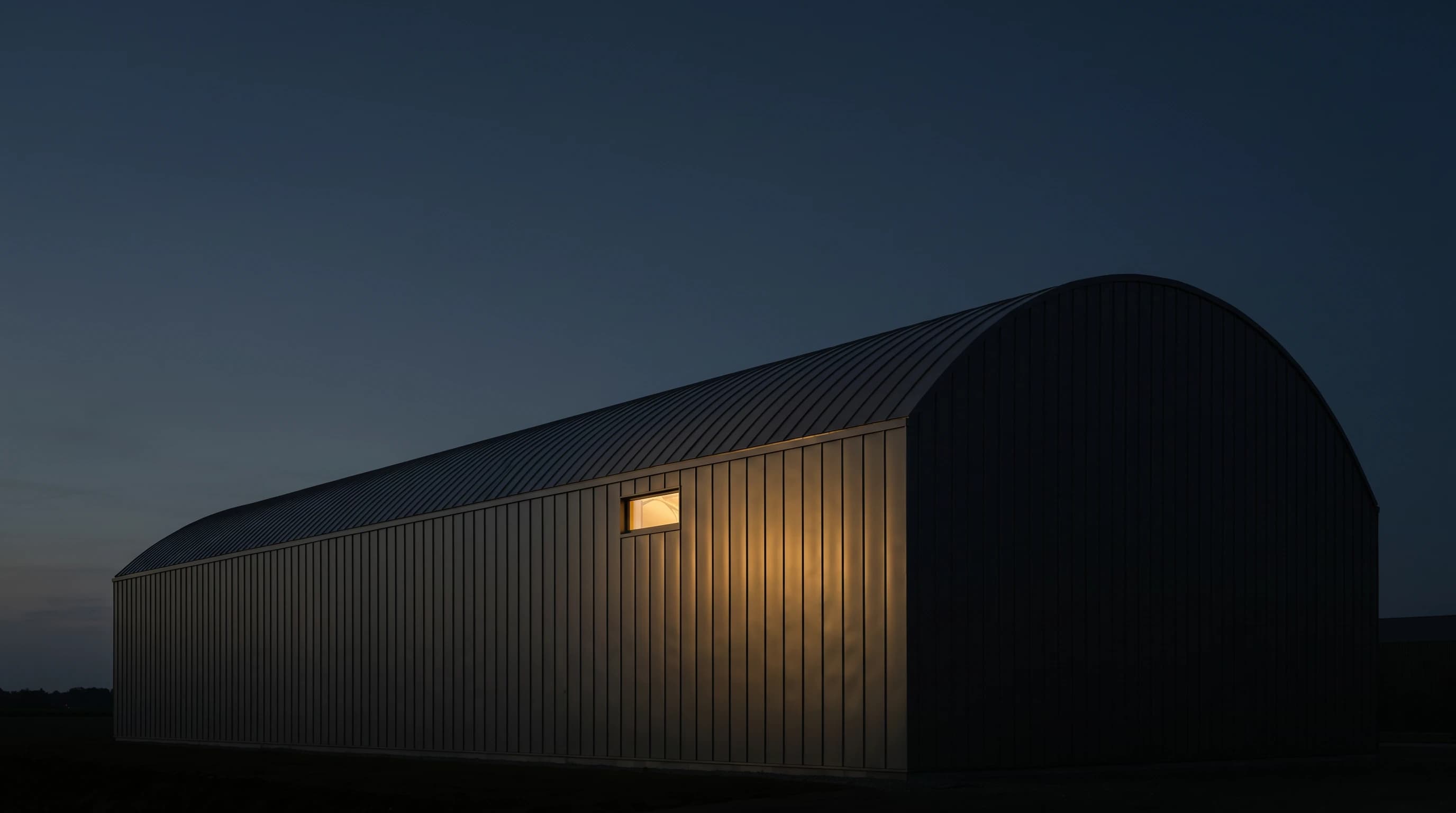 Long-span industrial building at dusk, entire envelope clad in matte aluminium standing-seam panels, one warm tungsten window glowing
