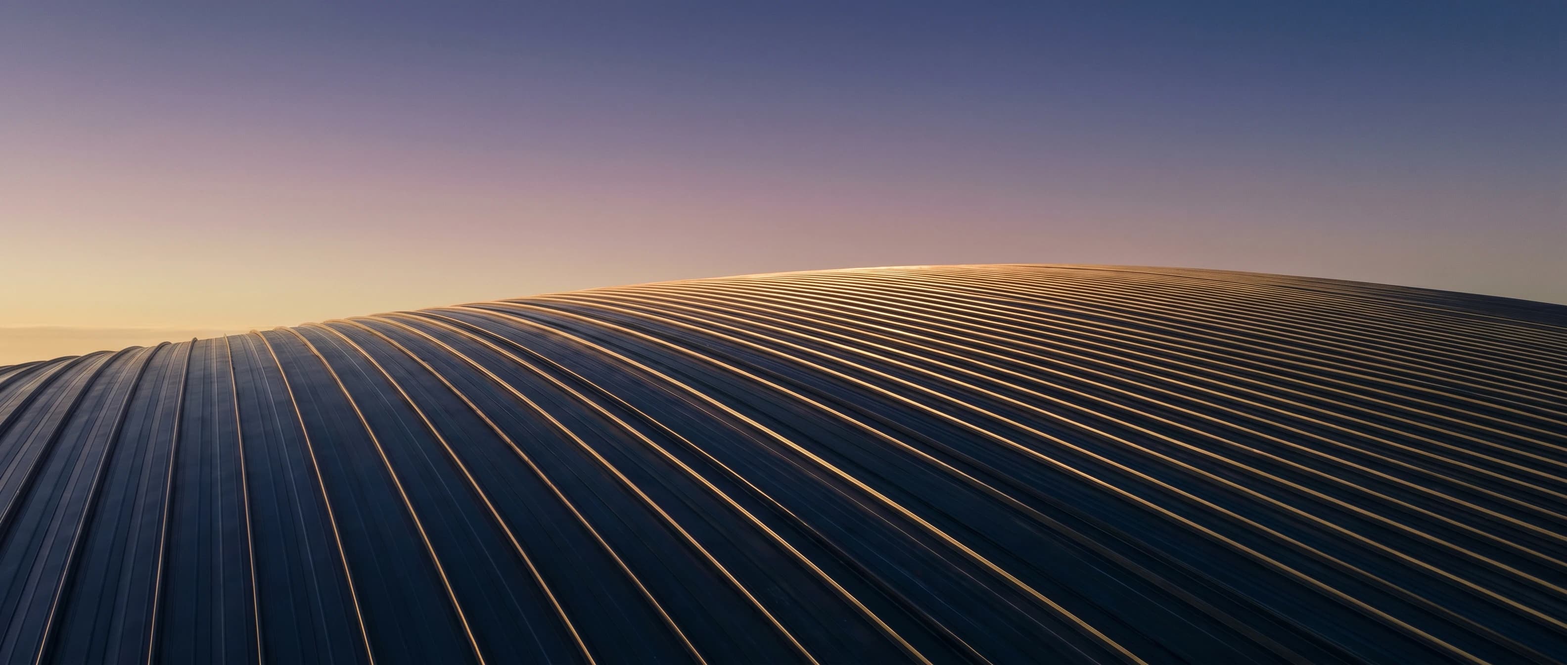 Curved standing-seam stadium roof at dawn, brass-warm seam highlights running across the metal surface